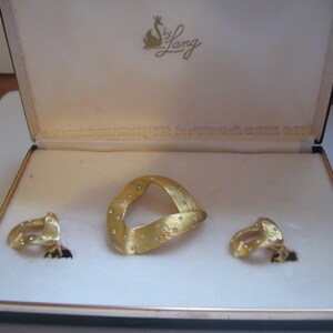 May include: A gold jewelry set in a white box with the text "by Lang" on the lid. The set includes a large, gold, diamond-encrusted pendant and two smaller, gold, diamond-encrusted earrings.