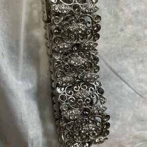 Pu&ograve; includere: Un braccialetto d'argento con un design delicato e ornato che presenta pi&ugrave; file di strass trasparenti e grigio scuro.