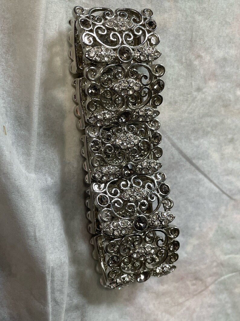 Pu&ograve; includere: Un braccialetto d'argento con un design delicato e ornato che presenta pi&ugrave; file di strass trasparenti e grigio scuro.