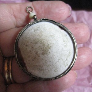 May include: A silver locket pendant with a white, round, textured surface. The locket has a small loop at the top for attaching a chain.