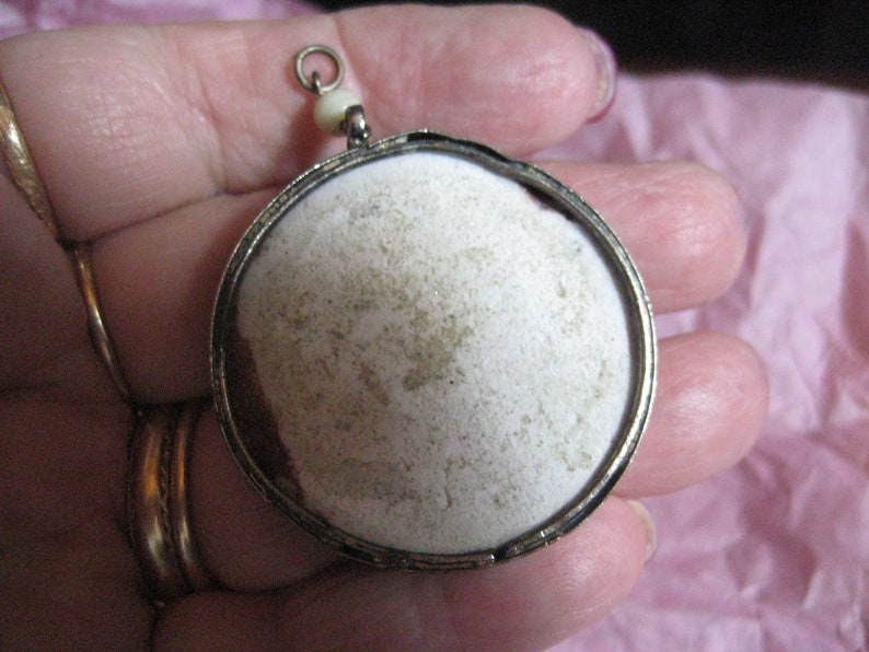 May include: A silver locket pendant with a white, round, textured surface. The locket has a small loop at the top for attaching a chain.