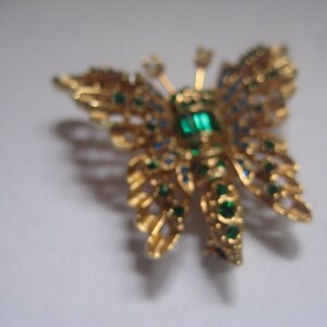 May include: A gold butterfly brooch with green and blue rhinestones. The butterfly has a detailed design and is a vintage style.