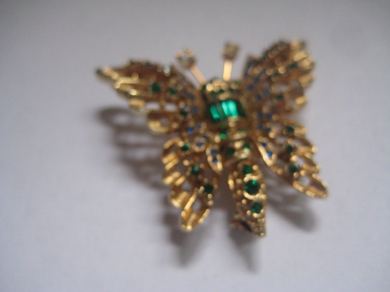 May include: A gold butterfly brooch with green and blue rhinestones. The butterfly has a detailed design and is a vintage style.