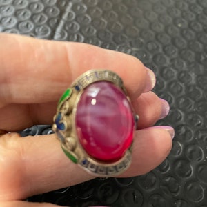 Puede incluir: Un anillo de plata con una piedra ovalada rosa en cabujón engastada en un engaste de plata. El anillo está decorado con esmalte verde y azul.