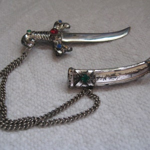 Könnte beinhalten: Silberfarbene Dolchbrosche mit gebogener Klinge und dekorativem Griff. Der Griff ist mit roten, blauen und grünen Edelsteinen verziert. Eine silberne Kette verbindet den Dolch mit seiner Scheide.