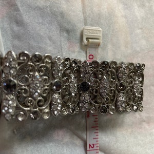 Pu&ograve; includere: Un braccialetto d'argento con un design a filigrana e strass trasparenti e neri. Il braccialetto ha un diametro di circa 5 cm.