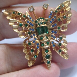 May include: A gold-tone butterfly brooch with green and blue rhinestones. The butterfly has a detailed design and is a vintage piece of jewelry.