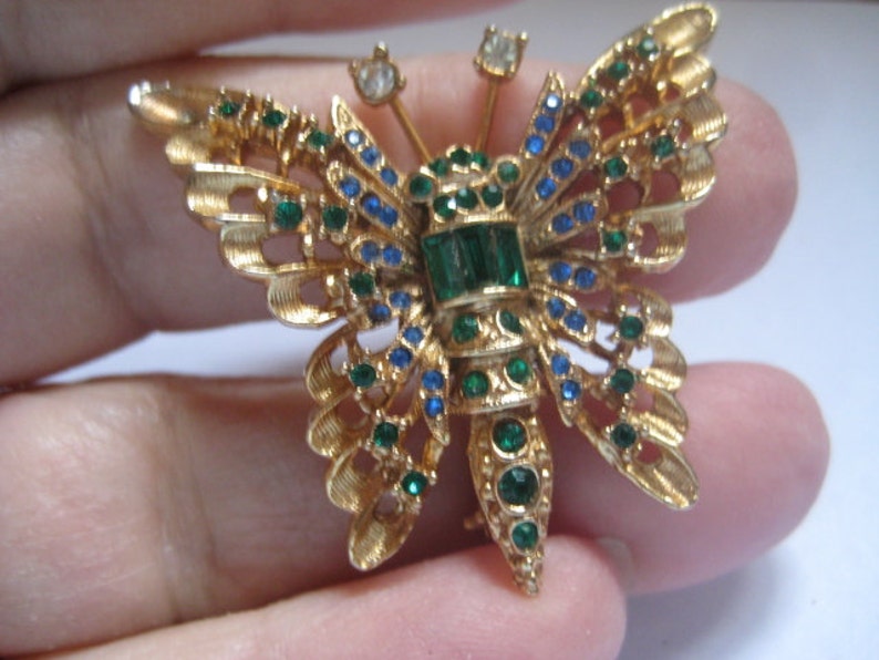 May include: A gold-tone butterfly brooch with green and blue rhinestones. The butterfly has a detailed design and is a vintage piece of jewelry.