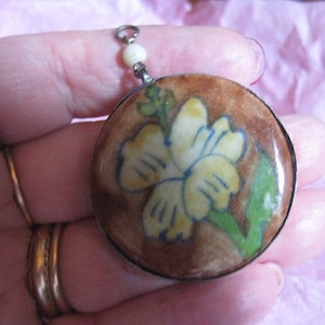 May include: A round pendant with a white flower with blue and yellow accents on a brown background. The pendant is attached to a silver chain with a white bead.