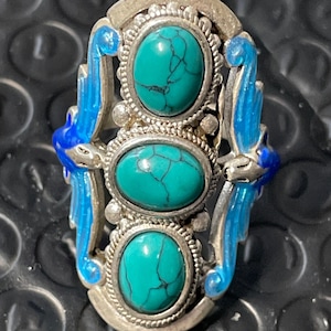 Puede incluir: Un anillo de plata con tres piedras turquesa engastadas en un diseño decorativo. El anillo presenta detalles de esmalte azul en los lados.