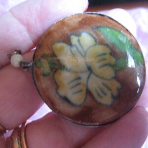 May include: A round, brown, ceramic pendant with a silver rim. The pendant features a yellow flower with green leaves painted on it.