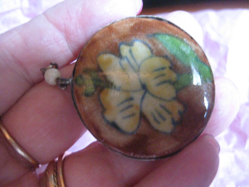 May include: A round, brown, ceramic pendant with a silver rim. The pendant features a yellow flower with green leaves painted on it.