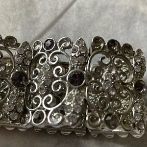 Pu&ograve; includere: Un braccialetto d'argento con un design a filigrana e strass trasparenti e viola.