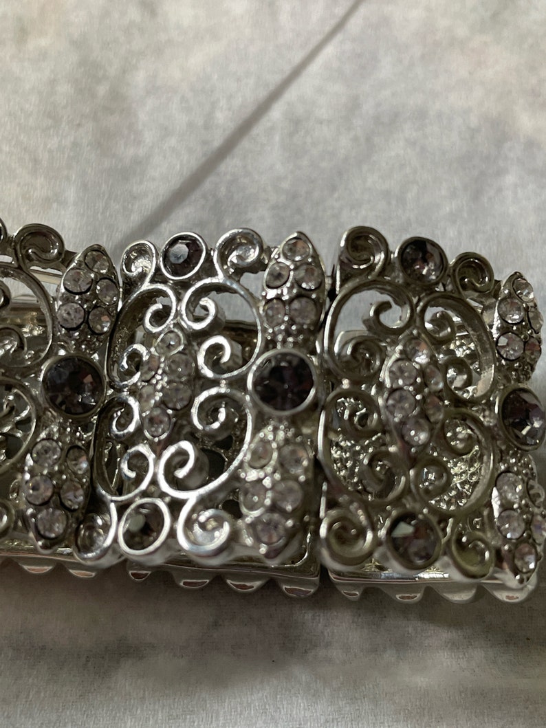 Pu&ograve; includere: Un braccialetto d'argento con un design a filigrana e strass trasparenti e viola.