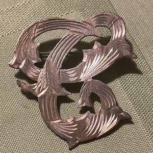May include: A silver-toned brooch with an abstract design. The brooch features a series of interconnected, stylized shapes with carved details. The design includes open spaces and curved lines, creating a flowing, organic aesthetic. The brooch is set against a textured green background.