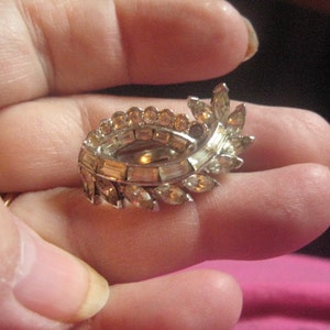 May include: Vintage silver-toned brooch with clear and amber-colored rhinestones. The brooch features an oval shape with a leaf-like design on one side. The rhinestones are set in a silver metal frame, creating a sparkling effect. A classic piece of jewelry.