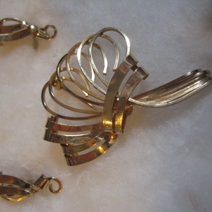May include: A set of gold-toned earrings and a brooch. The earrings are simple, with a curved shape. The brooch is more elaborate, with a fan-like design.