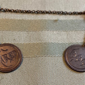 May include: Vintage silver-coloured collar pin featuring two coin-shaped pendants. Each pendant is linked to a pin with a chain. The pendants have engraved designs and lettering. The pins have a gold tone.
