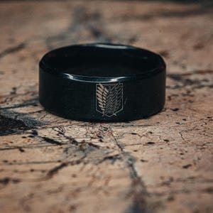 Op de afbeelding: Een zwarte ring met een matte afwerking. De ring heeft een gedetailleerd embleem op de voorkant. Het embleem is een gestileerd ontwerp met vleugels en een wapen. De ring ligt op een getextureerd, bruin oppervlak.