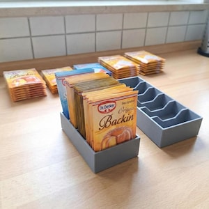 May include: Grey plastic organisers holding packets of baking powder and other ingredients. The packets are orange, blue, and white. The text "Dr. Oetker Backin" is visible on the packets. The organisers are on a light-coloured wooden surface.