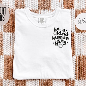 May include: White t-shirt with the phrase "be a kind human being" in black script. The shirt is folded and placed on a woven brown and white surface. The words "Comfort Colors" and "White" are in the image.