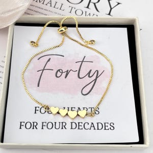 Op de afbeelding: Een gouden kettingarmband met een schuifsluiting en vier kleine gouden hartbedels. De armband wordt gepresenteerd in een witte doos met een roze achtergrond en de tekst "Forty" en "Four Hearts for Four Decades".