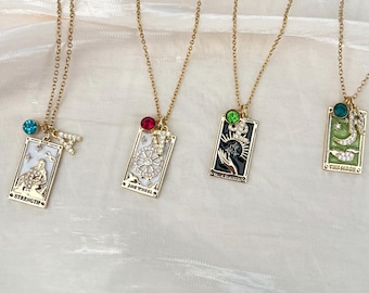 May include: Four gold-tone necklaces featuring rectangular tarot card pendants. The pendants depict Strength, The Wheel, The Magician, and The Moon with enamel details, small crystal accents, and attached letter charms with a single colored crystal.