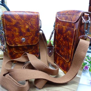 Peut inclure: Deux sacs rectangulaires verticaux avec une finition texturée marron et orange tachetée. Chaque sac présente un rabat avant avec une fermeture à pression en métal argenté et une large bandoulière en tissu tissé beige avec des mousquetons.