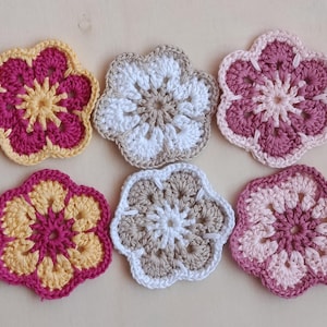 Könnte beinhalten: Sechs handgefertigte gehäkelte Blumenuntersetzer in Rosa-, Gelb- und Weißtönen. Die Untersetzer haben eine Blumenform mit einem detaillierten, durchbrochenen Design. Die Untersetzer sind auf einer hellen Oberfläche angeordnet.