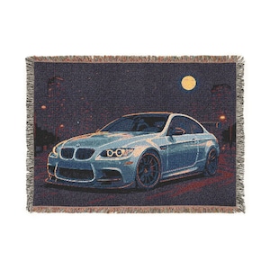 May include: Woven throw blanket featuring a light blue sports car with black wheels, set against a night scene with a full moon and city lights. The blanket has a fringed edge and a dark background.