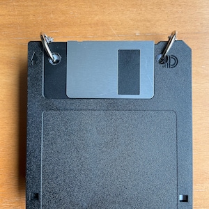 May include: A black 3.5-inch floppy disk with a silver metal slider and two silver key rings attached. The disk is on a wooden surface. The disk has a rectangular cutout and a small arrow symbol.