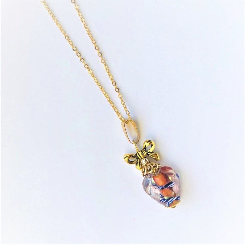 May include: A gold chain necklace with a heart-shaped pendant. The pendant is made of clear glass with orange and blue flecks and a gold bow on top.