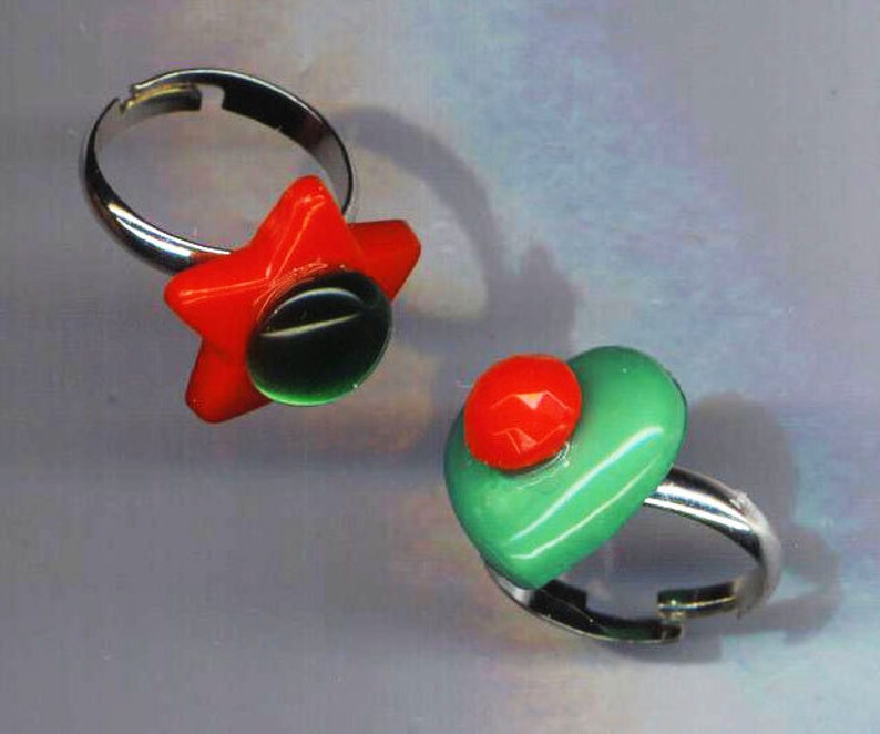 May include: Two silver rings with colorful glass accents. One ring features a red star with a green center. The other ring has a green heart with a red center.