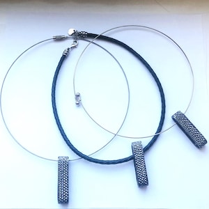 May include: Three necklaces with pendant bars. Two have silver-tone wire choker-style chains, and one has a dark blue braided leather cord. The pendant bars are dark gray with a textured surface. The necklaces are displayed on a white background.