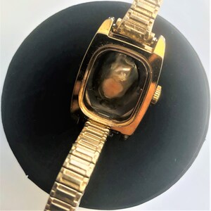 May include: A gold-toned wristwatch with a square face and a gold metal band. The watch face is missing and the watch is lying on a black surface.
