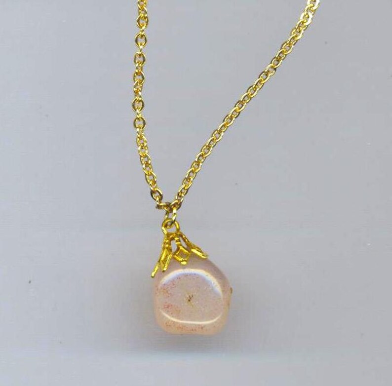 Puede incluir: Un collar de cadena dorada con un colgante de piedra rosa iridiscente. El colgante est&aacute; engastado en un marco de filigrana dorada.