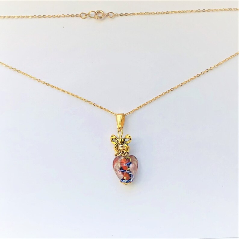 May include: A gold chain necklace with a heart-shaped pendant. The pendant is made of glass with a multi-colored pattern and a gold bow at the top.