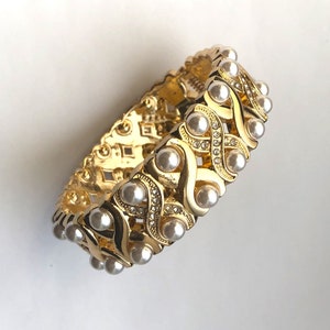 May include: A gold bracelet with a delicate, interwoven design accented with white pearls. The bracelet is flexible and has a polished finish.
