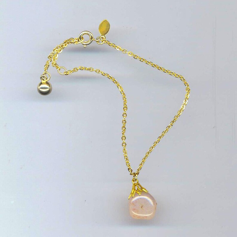 Puede incluir: Un brazalete de cadena dorada con un colgante rosa de forma irregular. El colgante est&aacute; unido a la cadena con un cierre dorado.