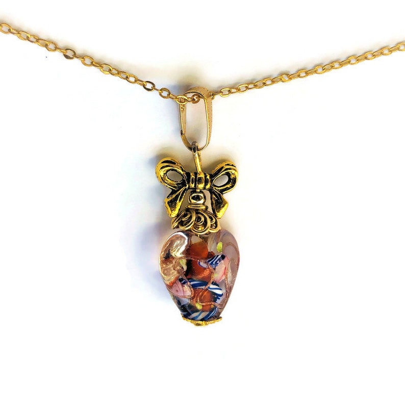 May include: A gold chain necklace with a heart-shaped pendant. The pendant is made of clear glass with colorful flecks and a gold bow on top.