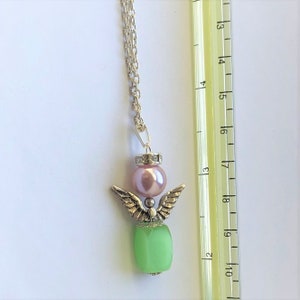 May include: A silver angel pendant necklace with a green glass bead, a pink pearl, and a silver chain. The pendant is approximately 1.75 inches long.