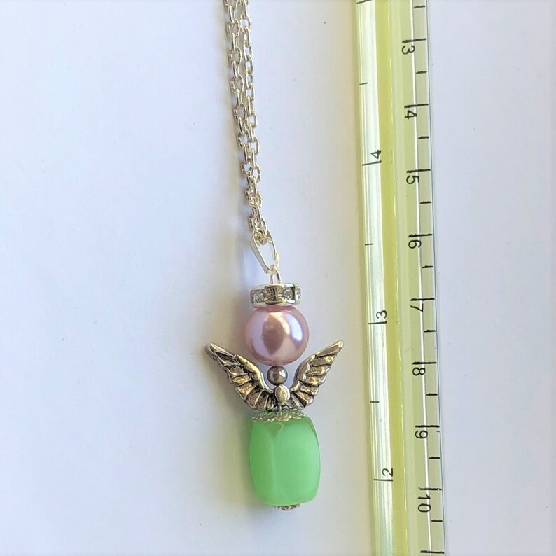 May include: A silver angel pendant necklace with a green glass bead, a pink pearl, and a silver chain. The pendant is approximately 1.75 inches long.