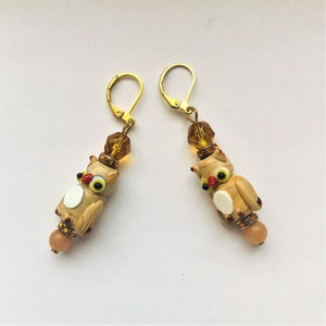 May include: A pair of owl-shaped earrings. The earrings feature a light brown owl design with yellow eyes, red accents, and a white belly. The earrings have gold-colored lever-back closures and are adorned with amber-colored beads.