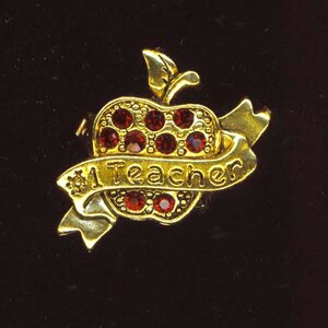 May include: A gold-tone pin in the shape of an apple with red rhinestones. The apple has a banner that reads "A Teacher".