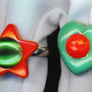 May include: Two colorful rings with a red and green design. The ring on the left is a star shape with a green center and red outer edges. The ring on the right is a heart shape with a green outer edge and a red center.
