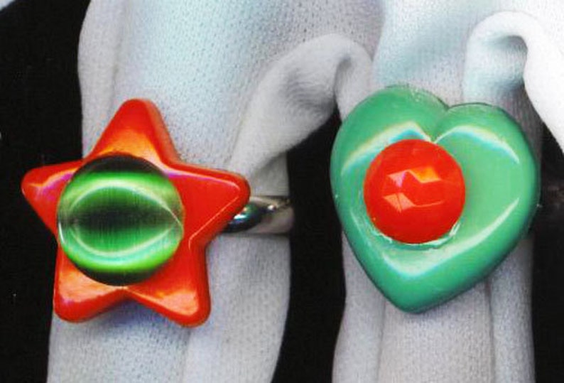 May include: Two colorful rings with a red and green design. The ring on the left is a star shape with a green center and red outer edges. The ring on the right is a heart shape with a green outer edge and a red center.