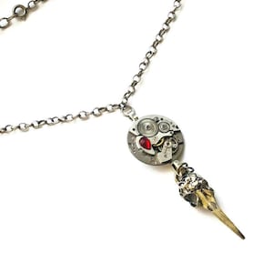 May include: A silver-toned necklace featuring a pendant with watch gears, a red gem, and a bird skull charm. The chain is a simple link design, and the pendant hangs from a small loop.