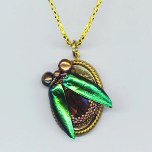 May include: A gold pendant necklace with a green beetle wing design. The beetle wings are iridescent and have a metallic sheen. The pendant is set in a gold oval frame and is surrounded by small beads.