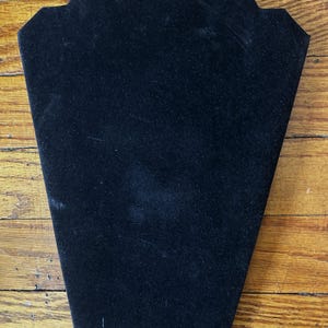 May include: A black velvet-covered necklace display stand featuring a tapered, shield-like shape with a flat top edge and angled sides designed to hold jewelry pieces securely for presentation purposes in a retail or personal collection setting.