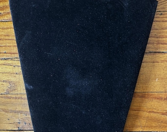 May include: A black velvet-covered necklace display stand featuring a tapered, shield-like shape with a flat top edge and angled sides designed to hold jewelry pieces securely for presentation purposes in a retail or personal collection setting.